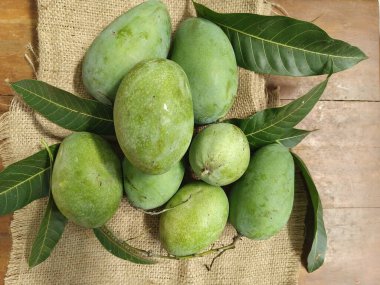 Fresh green mangoes with leaves arranged on burlap fabric, showcasing tropical fruit in a natural rustic setting.
