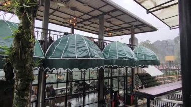 Endüstriyel tasarımı, cam pencereleri, yeşil tenteleri, asılı ışıkları ve yemyeşil yağmurlu çevresi olan modern çok katlı kafe, Malang, Endonezya - 09 Şubat 2026