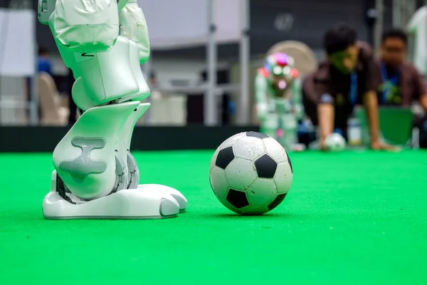 Futbol robotunun sağ bacakları suni futbol sahasında futbol oynarken, arka planda bulanık insanlar... 