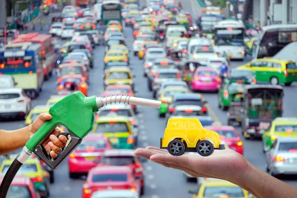 Sağdaki sarı oyuncak arabanın benzinini doldurmak için otomatik hortum tutan adamların sol kolları bulanık arka planda trafik sıkışıklığı, endüstriyel yağ ve gaz enerjisi kavramı