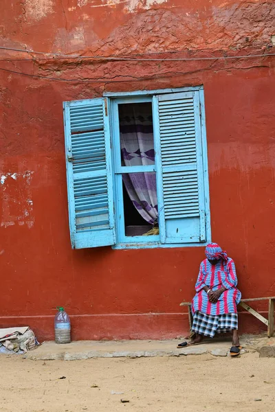 28-08-2025 Goree Adası Senegal: Kırmızı koloni duvarında mavi pencerenin yanında dinlenen renkli elbiseli yaşlı kişi