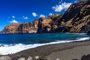 Stunning landscape of Los Gigantes cliffs on Tenerife, Canary Islands, with cactus growing on a rocky slope above the deep blue Atlantic Ocean under a partly cloudy sky. Travel, nature, and adventure concept.