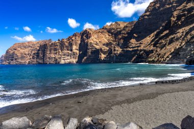 Stunning landscape of Los Gigantes cliffs on Tenerife, Canary Islands, with cactus growing on a rocky slope above the deep blue Atlantic Ocean under a partly cloudy sky. Travel, nature, and adventure concept.