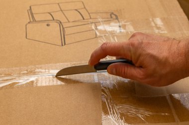 Karton bir kutuyu açmak için bıçak kullanan bir adamın eli bantla kapatılmış. Pakette bir kanepenin basılı bir illüstrasyonu yer alıyor ve yeni mobilyaların açılması veya eve teslim edilmesi öneriliyor. Kutu açma, nakliye, teslimat ve inte kavramı