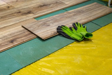 Üzerinde yeşil ve siyah koruyucu eldivenler bulunan ahşap kaplamalı döşeme panellerini kapatın. Ev yenileme, döşeme kurulumu ve DIY doğal ışık iyileştirme kavramı.