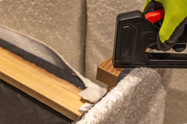 İşçiler, ev atölyesindeki ahşap döşemelere ya da mobilya yenileme çalışmalarına hafif kumaş eklemek için siyah zımba tabancası kullanarak yeşil koruyucu eldivenler veriyorlar.