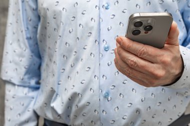 Uzun kollu mavi gömlekli ve şık pantolonlu bir adam hanında cep telefonu olan beton bir duvarın karşısında duruyor.