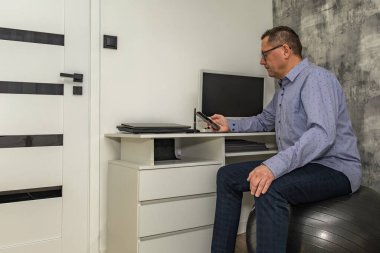 Mavi gömlekli erkek ofis çalışanı bilgisayarın önünde ortopedik topun üzerinde oturuyor, evden uzaktan çalışırken işyeri organizasyonu, ofis çalışanları sırt problemi