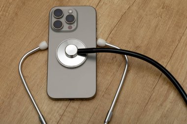 Hasarlı bir cep telefonu ahşap servis masasına yerleştirilmiş, steteskop kulaklıkları, telefon arızası aramak, telefonu kendin tamir etmeye çalışmak, cep telefonu servisi.