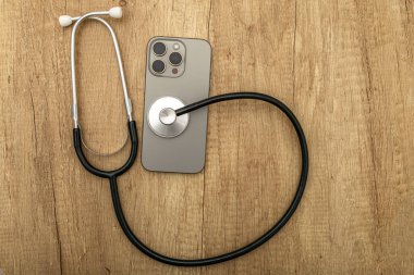 Hasarlı bir cep telefonu ahşap servis masasına yerleştirilmiş, steteskop kulaklıkları, telefon arızası aramak, telefonu kendin tamir etmeye çalışmak, cep telefonu servisi.