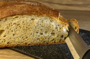 Ekmek kesmek için keskin bıçaklı bir bıçak, mutfak tezgahında bir somun taze ekmek dilimlemek, ekmek kesme tahtası, dilimlenmiş ekmek, ahşap tezgahta bir dilim ekmek.
