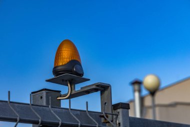 Kapı otomatik olarak kapanır. Uzaktan kumandayla, çitlere takılı turuncu bir uyarı lambası, giriş kapısında trafik ışıkları, ve çitin üzerinde turuncu bir lamba..