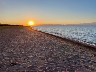Issyk-Kul 'daki el değmemiş kumlu plajın arka planında kar kaplı Tian Shan dağları gün batımında, Kırgızistan. Yüksek kalite fotoğraf