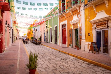 Campeche, Meksika. Renkli binaları ve kırmızı tencerede yeşil bitkileri olan bir cadde. Sokaklar masa ve sandalyelerle dolu ve etrafta yürüyen insanlar var.