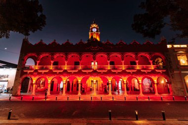 Merida, Yucatan, Meksika, kırmızı kemerli büyük bir bina ve saat kulesi. Bina kırmızıyla aydınlatılmış, sıcak ve davetkar bir atmosfer sağlıyor.