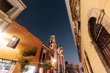 Merida, Yucatan, Meksika, geceleri arka planda yüksek bir bina olan sokak sahnesi. Sokaklar bina ve ağaçlarla dolu ve etrafta dolaşan bir sürü insan var.