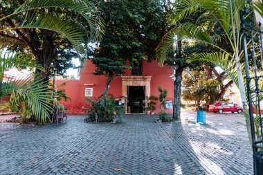 Merida, Yucatan, Meksika, beyaz kapılı kırmızı bina yemyeşil bir avlunun önünde duruyor. Avlu ağaçlar ve bitkilerle dolu. Huzurlu ve davetkar bir ortam yaratıyor. Park yerinde bir araba var.