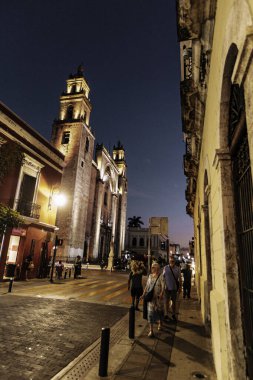 Merida, Yucatan, Meksika, bir grup insan geceleri arka planda büyük bir binayla caddede yürüyor. Sahne huzurlu ve huzurlu, insanlar yürüyüşlerinden ve sessiz atmosferden hoşlanıyor gibi görünüyor.