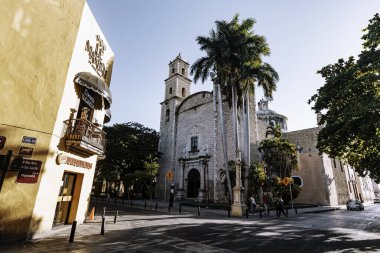 Merida, Yucatan, Meksika, sarı binalı ve kiliseli sokak manzarası. Sokakta yürüyen insanlar ve park etmiş bir araba var.