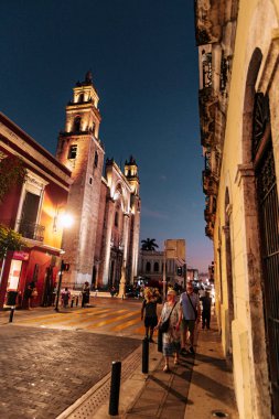 Merida, Yucatan, Meksika, bir grup insan geceleri arka planda büyük bir binayla caddede yürüyor. Sahne huzurlu ve huzurlu, insanlar yürüyüşlerinden ve sessiz atmosferden hoşlanıyor gibi görünüyor.