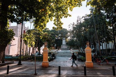 Merida, Yucatan, Meksika, heykeli ve bankı olan bir park. Parkta bir adam yürüyor. Birkaç bank ve üzerinde oturan birkaç kişi var.