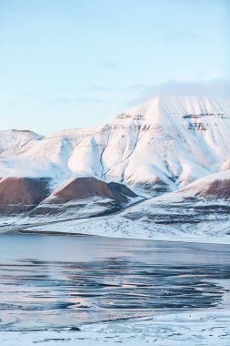 Longyerbeyen, Svalbard, dağ sıraları karla kaplı ve ön planda bir göl. Gökyüzü mavi ve dağlar arka planda.