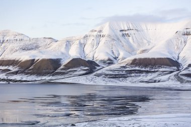 Longyerbeyen, Svalbard, önünde göl olan Karlı dağ sıraları. Gökyüzü açık ve güneş parlıyor. Huzurlu ve huzurlu bir atmosfer yaratıyor.