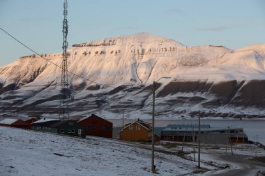 Longyerbeyen, Svalbard, birkaç ev uzaklıktaki Karlı dağ sıraları. Evler küçük ve kırmızı ve suda bir tekne var. Huzurlu ve huzurlu bir manzara, karla kaplı bir manzara ve sakin bir su