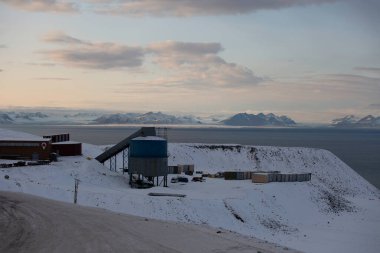 Longyerbeyen, Svalbard, büyük mavi tank karlı yamaçta bir su kütlesinin yanında oturur. Gökyüzü bulutlu ve güneş batıyor.