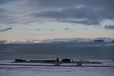 Longyerbeyen, Svalbard, arka planda büyük bir su kütlesi olan karlı bir manzara. Gökyüzü bulutlu ve güneş batıyor.