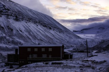 Longyerbeyen, Svalbard, Kırmızı bina karlı bir yamaçta. Gökyüzü pembe ve turuncu renklerin bir karışımıdır. Huzurlu ve huzurlu bir atmosfer yaratır. Bina bir kulübe gibi görünüyor ve çevresi karla kaplı.