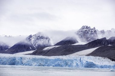 Svalbard seyahati. Önünde mavi bir buzulla dağ sırası. Gökyüzü bulutlu ve dağlar karla kaplı.