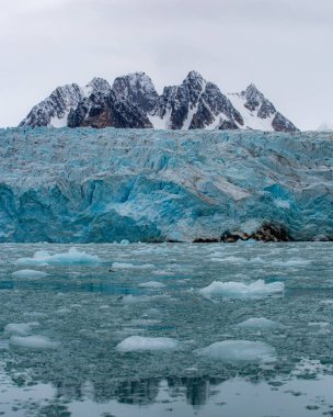 Svalbard seyahati - Büyük dağ sıraları suya yansıyor. Gökyüzü bulutlu ve su donmuş.