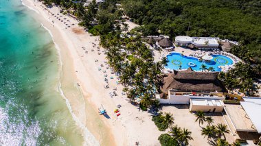 Arka planda tatil köyü olan bir plaj. Otelin bir havuzu ve birkaç kabini var.