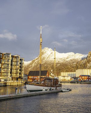 Svolvear, Lofoten Adası, Norveç küçük teknesi büyük bir binanın önündeki rıhtıma yanaştı. Gökyüzü bulutlu ve güneş batıyor.