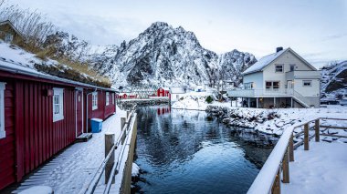 Svolvear, Lofoten Adası, Norveç Karlı manzarası bir nehir ve kırmızı evler sırası. Evler ahşaptan yapılmış ve kırsal bir görünüme sahipler. Nehir sakin ve çevresindeki manzarayı yansıtıyor.