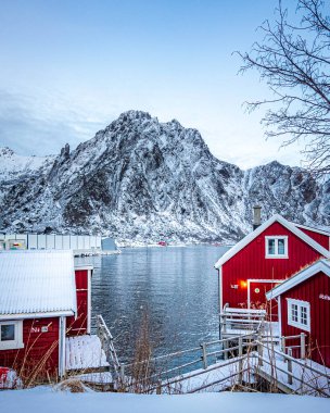 Svolver Norveç Lofoten Adaları Kırmızı evi karlı bir yamaçta, bir su kütlesinin yanında duruyor. Sahne sakin ve sakin, arka planda dağlar ve batan güneşin ışığını yansıtan su. Evler küçük ve rahat.