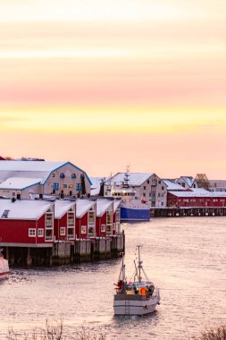 Svolver Norveç Lofoten Adaları Teknesi kırmızı binaların bulunduğu bir kasabanın yakınındaki bir nehirde yol alıyor. Gökyüzü turuncu ve pembe. Sıcak ve huzurlu bir atmosfer yaratıyor.