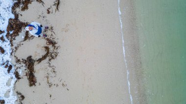 Beyaz şapkalı bir adam plajda duruyor. Kumsal yosunla kaplı ve su yeşil.