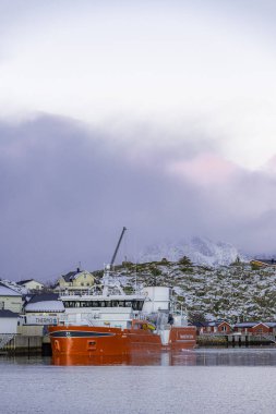 Kırmızı tekne bir dağın yanındaki suyun içinde. Gökyüzü bulutlu ve güneş batıyor.