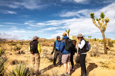 Joshua Tree Ulusal Park Grubu bir çölde duruyor, bazıları sırt çantası takıyor. Birbirleriyle konuşuyorlar ve birlikte iyi vakit geçiriyorlar gibi görünüyor.