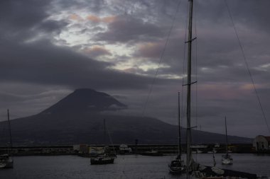 Bir grup yelkenli, arka planda bir dağ bulunan bir limana yanaştı. Gökyüzü bulutlu ve güneş batıyor, kasvetli bir atmosfer yaratıyor Faial Azores Portugal