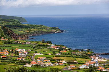 Faial, Azores Adaları, Portekiz. Arka planda mavi bir okyanus olan küçük bir kasabanın güzel manzarası. Evler beyaz ve kırmızı, çimenler yeşil. Sahne huzurlu ve sakin, okyanus rahatlatıcı bir zemin sağlıyor.
