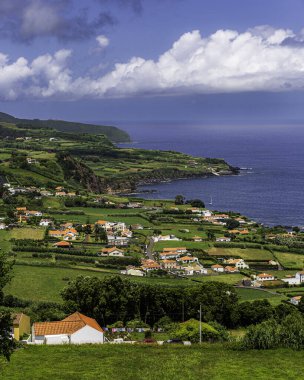 Faial, Azores Adaları, Portekiz. Arka planda plajı olan küçük bir kasabanın güzel manzarası. Evler küçük ve beyazdır ve kasaba yeşil tarlalarla çevrilidir. Gökyüzü mavi ve güneş parlıyor. Huzurlu ve huzurlu bir atmosfer yaratıyor.