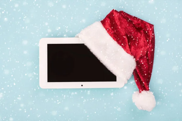 Christmas concept background.Digital tablet in santa claus hat with snowflakes over blue background