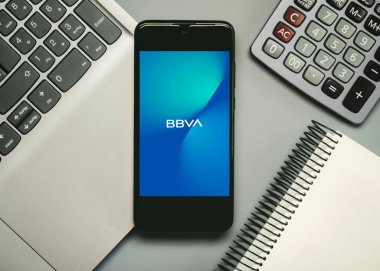 Akıllı telefon ekranında BBVA mobil uygulama logosu. Gri bir grupta dizüstü bilgisayar ve hesap makinesi yanında.