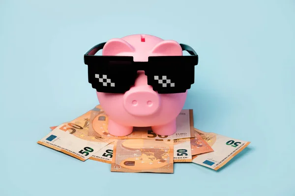 Pink piggy money bank with black sunglasses and euro bills on blue background