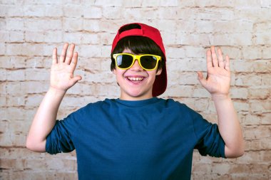 Şapkalı ve güneş gözlüklü mutlu ve gülümseyen çocuk avuçlarını beyaz tuğla duvarda gösteriyor.