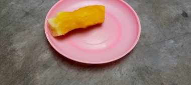 Canlı sarı, soyulmuş bir mango parçası küçük pembe bir plaka üzerinde, yüksek açıyla çekilmiş. Taze, tropikal meyve çerezi.