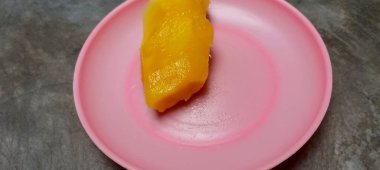 Canlı sarı, soyulmuş bir mango parçası küçük pembe bir plaka üzerinde, yüksek açıyla çekilmiş. Taze, tropikal meyve çerezi.